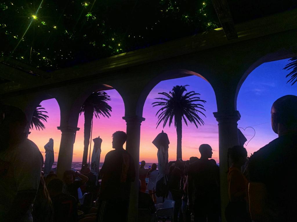 Colorful sunset looking out towards the beach with silhouettes of palm trees and cafe goers. The ceiling is scattered with green lights that look like stars.  